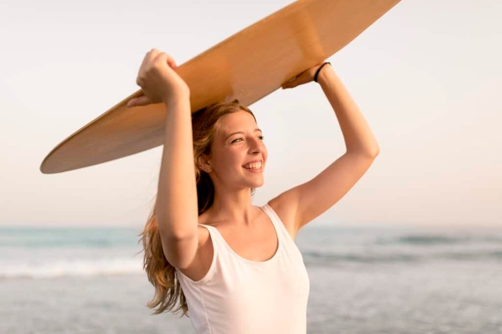 Mujer sonriente sosteniendo una tabla de surf sobre su cabeza en la playa, con el mar y el cielo de fondo.