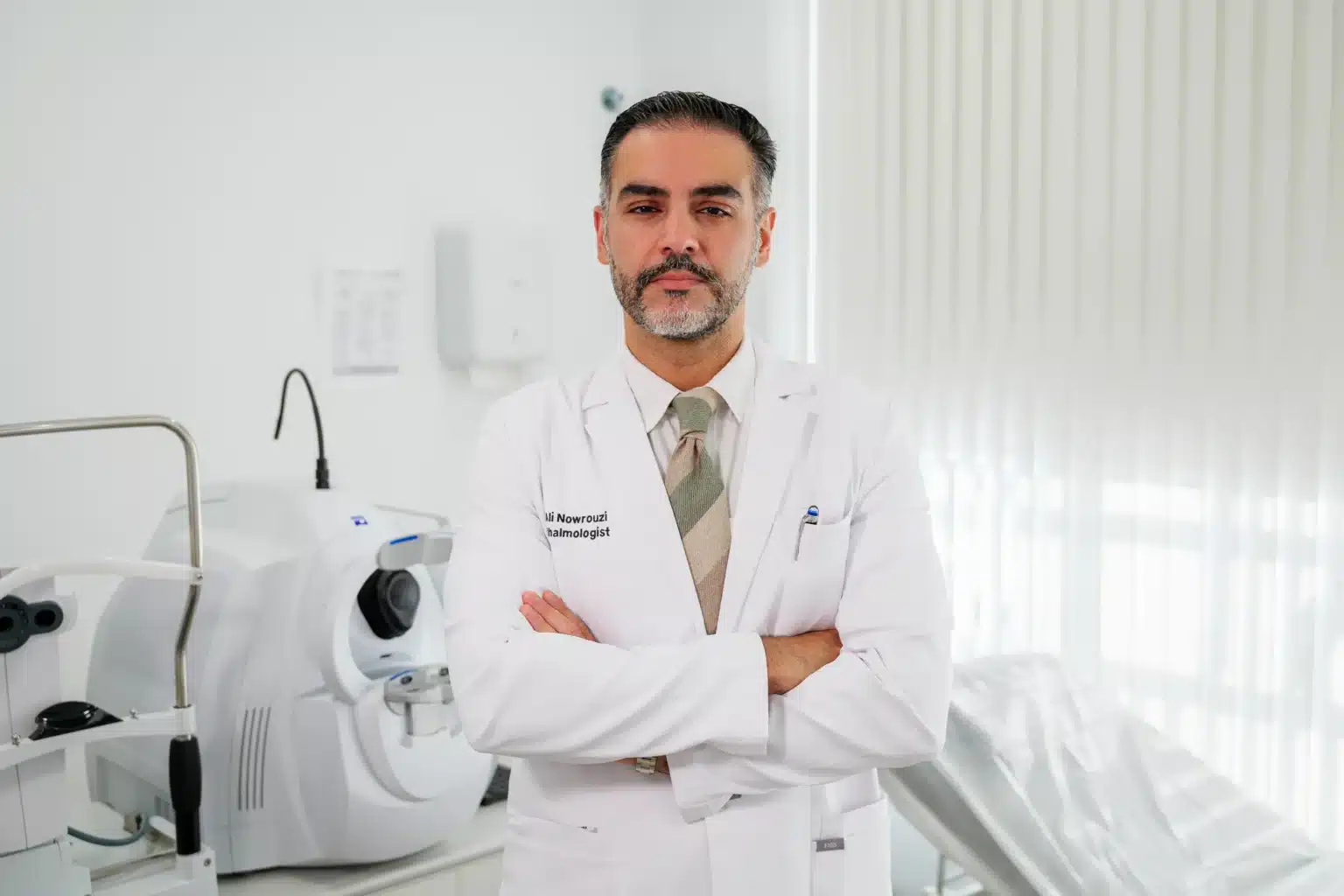 Médecin dans un cabinet dentaire, vêtu d'une blouse blanche, les bras croisés, avec du matériel dentaire en arrière-plan.