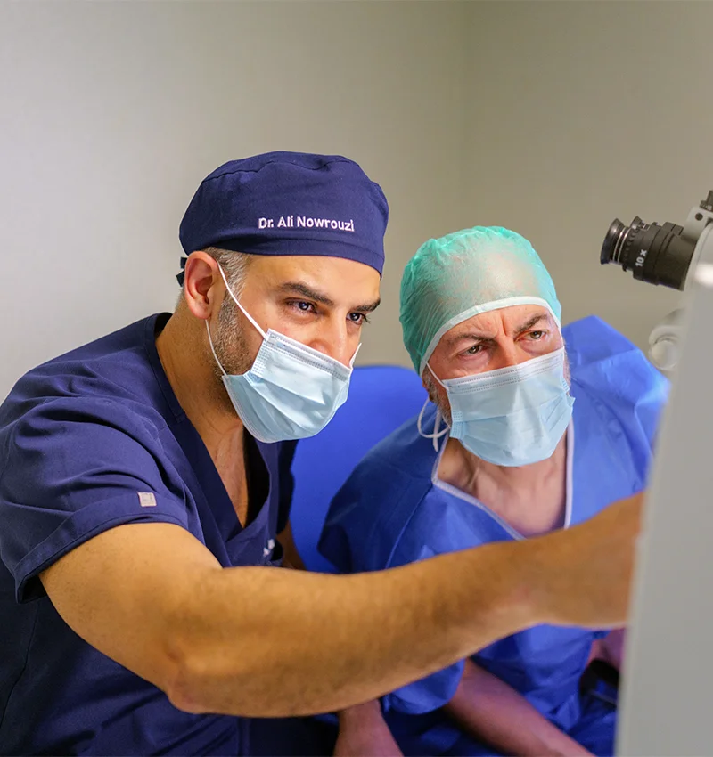 Dr. Ali Nowrouzi and a colleague, one in a blue cap and gown and the other in a green cap, look intently at a screen during a medical consultation, both wearing face masks.