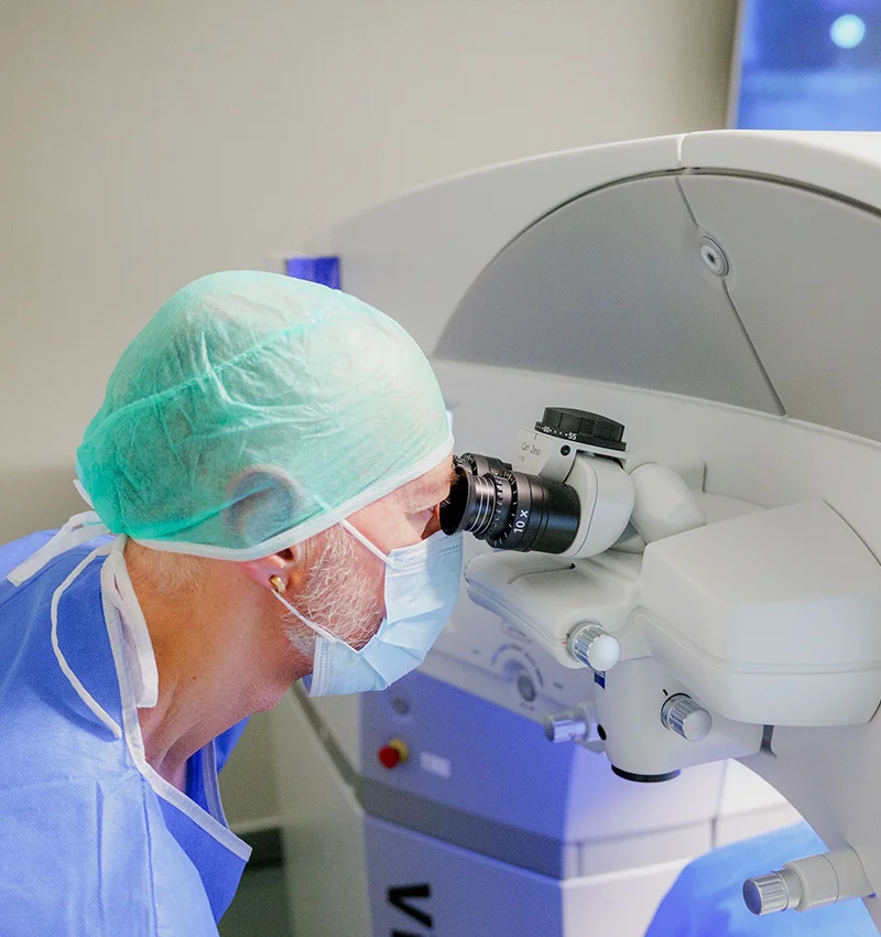 Physician using a microscope in a clinical setting, wearing a cap and mask, focusing on a surgical procedure.