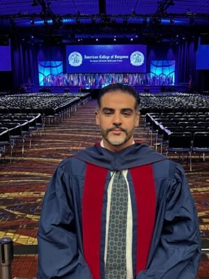 Oftalmólogo en Marbella 33 Hombre graduado con toga y birrete posando frente al escenario de una ceremonia de graduación en el Southern College of Optometry. Fondo de asientos vacíos y decoraciones del evento.